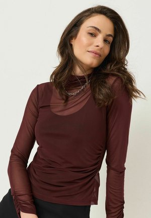 Femme aux longs cheveux bruns portant un haut transparent bordeaux à manches longues et un collier en chaîne argentée, posant devant un fond clair uni.