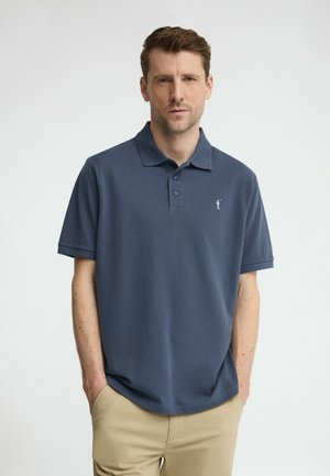 Hombre con camisa polo de manga corta azul marino y pantalones beige, de pie con las manos en los bolsillos contra un fondo claro y sencillo.