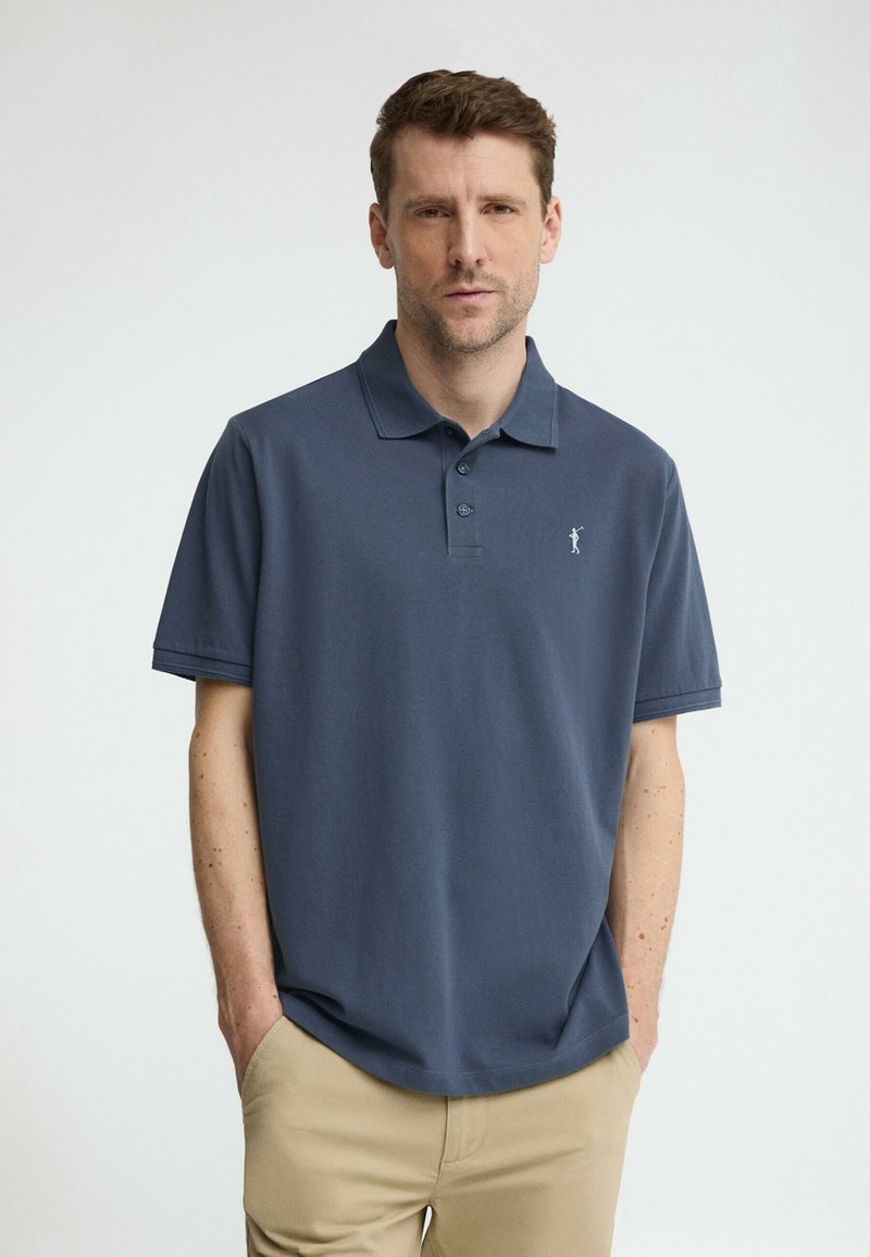 Uomo che indossa una polo a maniche corte blu scuro e pantaloni beige, in piedi con le mani nelle tasche contro uno sfondo chiaro e uniforme.