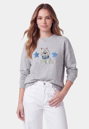Kvinde bærer grå sweatshirt med blå stjerner og hvid hundeillustration, kombineret med hvide bukser og blå hoop-øreringe.