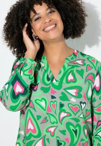 Grüne Bluse mit einem Herzmuster in verschiedenen Rosa- und Weißtönen, mit V-Ausschnitt und langen Ärmeln. Das Material wirkt glatt und leicht.