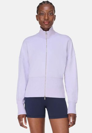 Vrouw draagt een lichtpaarse trui met rits en hoge kraag en donkerblauwe aangesloten korte broek, staande tegen een witte achtergrond.