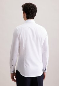 Camicia bianca con bottoni, maniche lunghe, texture liscia, dettagli con bottoni neri sui polsini e orlo curvo. La vista posteriore mostra il colletto e la vestibilità.