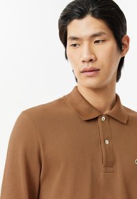 Brun polo tröja med en texturerad tyg, med krage, två knappar fram och en liten logotyp på vänster sida.