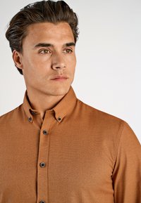 Brązowa koszula z guzikami, wykonana z fakturowanej tkaniny, z wyrazistym kołnierzykiem i czarnymi guzikami. Koszula jest noszona przez mężczyznę o włosach średniej długości.