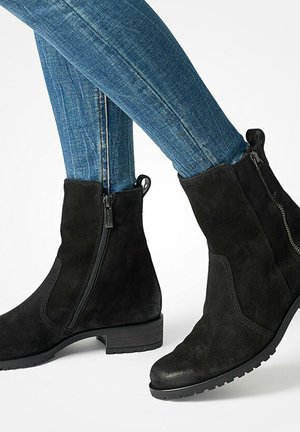 Schwarze Wildleder-Stiefeletten mit seitlichen Reißverschlüssen, getragen mit blauen Skinny-Jeans auf weißem Hintergrund, die die Sohle und den niedrigen Absatz der Stiefel zeigen.