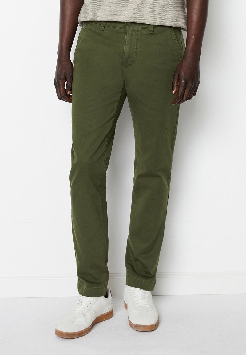 Homme portant un pantalon vert olive et des baskets blanches, debout contre un mur blanc, les mains détendues sur les côtés.
