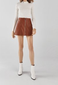 Hnědá mini sukně z umělé kůže s předním zipem, ve tvaru A a bočními kapsami. V kombinaci s bílým přiléhavým dlouhým rukávem a bílými kotníkovými botami.