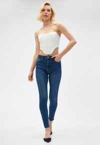 Top blanco sin mangas con diseño estructurado combinado con jeans ajustados de un azul oscuro. La modelo lleva zapatos planos negros de punta. Texturas simples y suaves.