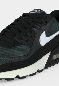 Baskets Nike Air Max noires et vert foncé avec logo Swoosh blanc et semelle épaisse blanche et noire, en gros plan.