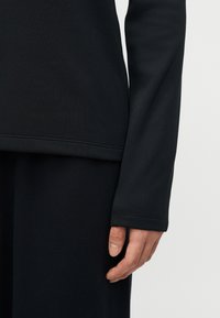 Δεξί χέρι και κάτω μπράτσο σε μακρυμάνικο ένδυμα μαύρου χρώματος, στηριγμένα δίπλα σε μαύρο παντελόνι πάνω σε απλό φωτεινό φόντο.