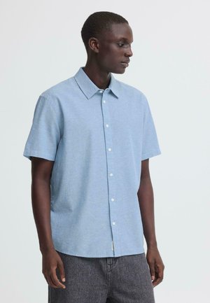 Młody mężczyzna w jasnoniebieskiej koszuli z krótkim rękawem na guziki i ciemnoszarych spodniach, patrzący w prawo na jednolitym tle.