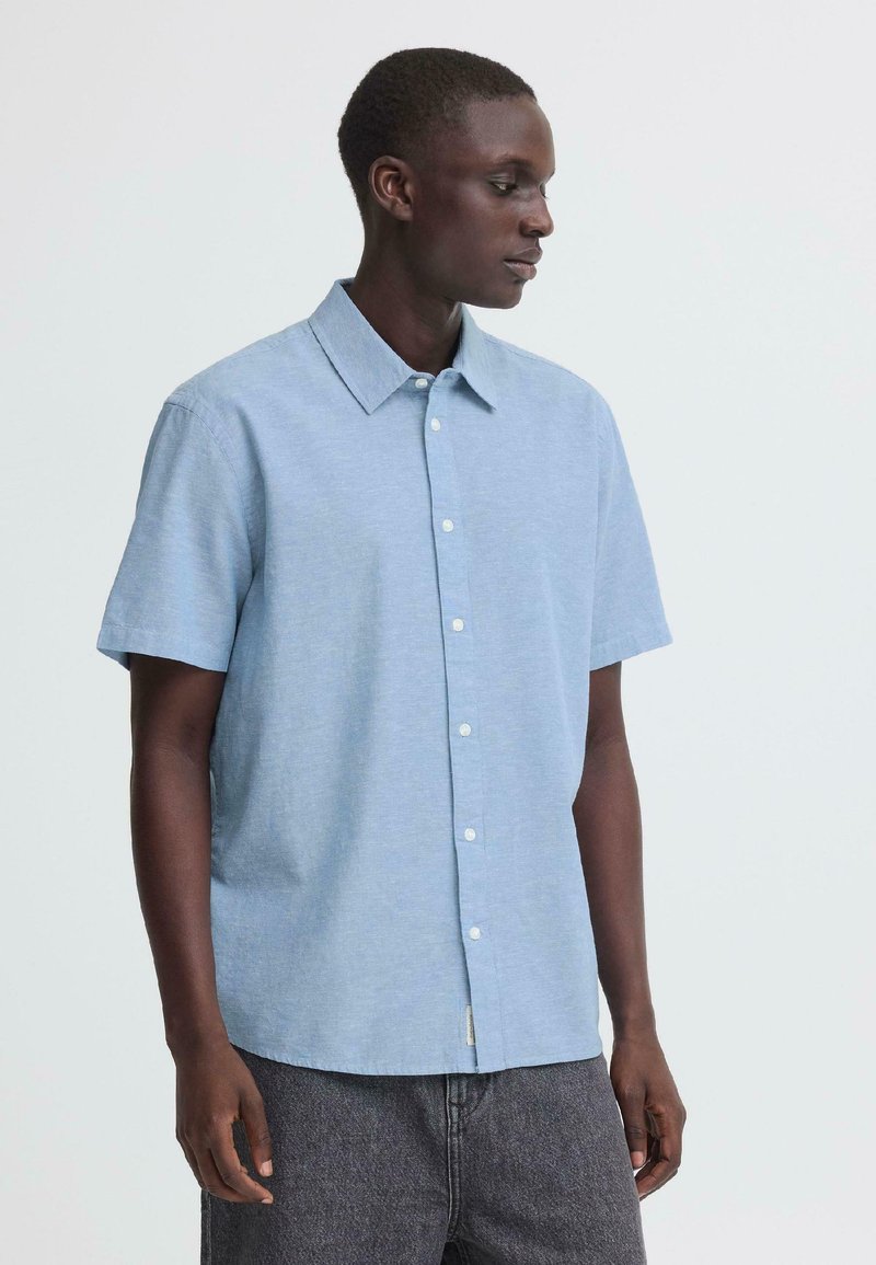 Νεαρός άνδρας με ανοιχτό μπλε κοντομάνικο πουκάμισο με κουμπιά και σκούρο γκρι παντελόνι, κοιτάζει προς τα δεξιά του σε απλό φόντο.