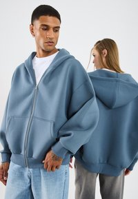 Hoodie in zachte blauwe stof met een rits aan de voorkant, oversized pasvorm en buidelzak. Heeft verlaagde schouders en geribbelde manchetten.