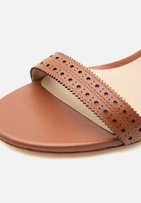 Καφέ δερμάτινο σανδάλι με φαρδιά διάτρητη λωρίδα πάνω από τα δάχτυλα και λεία σόλα, απεικονιζόμενο σε κοντινή λεπτομέρεια.