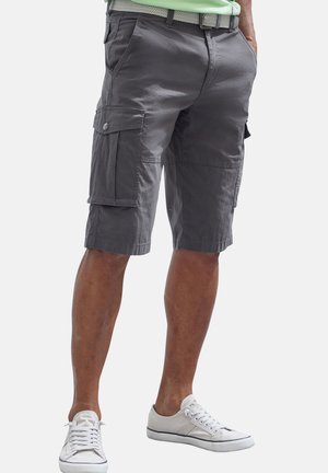 Man wearing dark gray cargo shorts with side pockets, a light belt, white sneakers, and a light green shirt with hands in pockets.