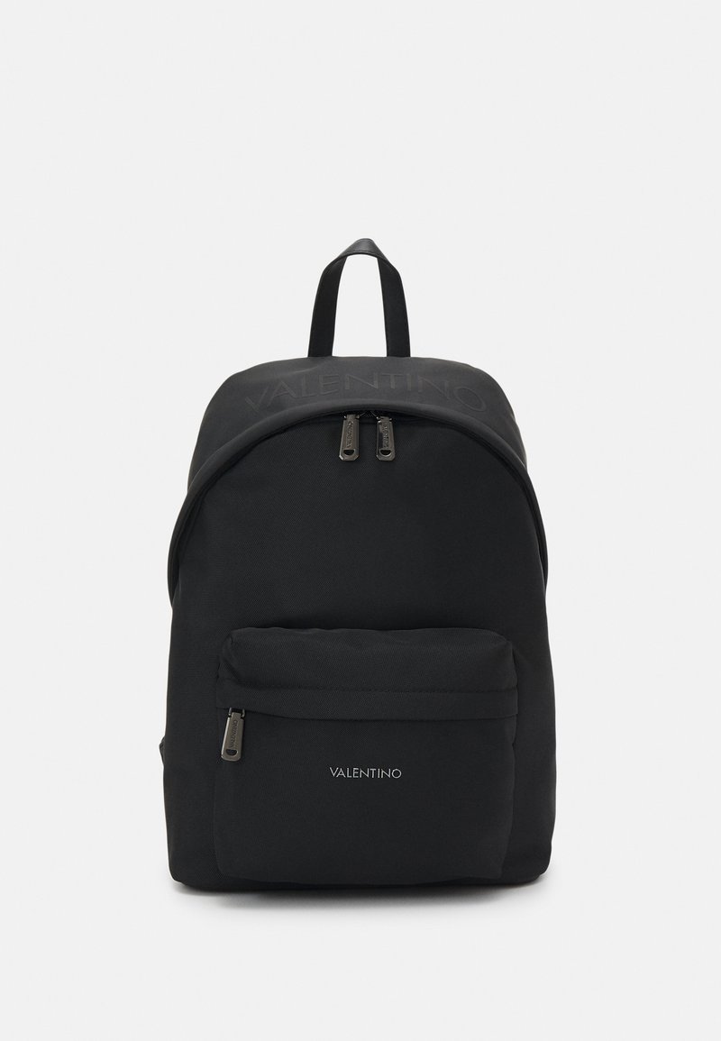 Mochila negra de nylon con parte superior redondeada, dos compartimentos con cremallera y bolsillo frontal. Presenta un sutil detalle del logotipo y una textura de tela resistente.