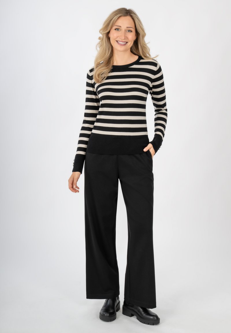 Gestreifter Pullover in Schwarz und Creme mit langen Ärmeln und einem schwarzen Bund, kombiniert mit schwarzen weiten Hosen und klobigen schwarzen Schuhen.