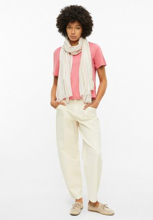 Femme aux cheveux bouclés porte un top rose clair à manches courtes, un pantalon large couleur crème, des chaussures beige et un foulard léger, debout les mains dans les poches.