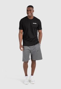 Sort kortærmet t-shirt med hvidt logo, parret med grå shorts og hvide sneakers. Casual atletisk tøj på en stående mandlig model.
