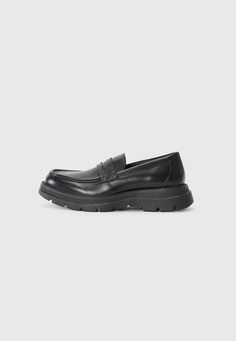 Mocassin en cuir noir avec une semelle épaisse à crampons, couture moc toe et une bride penny sur le devant, présenté en profil latéral sur fond blanc.