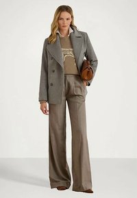 Γυναίκα που στέκεται φορώντας ένα γκρι διπλόχρωμο παλτό, καρό καφέ πουλόβερ και φαρδιά παντελόνια, κρατώντας μία καφέ τσάντα στον βραχίονα.