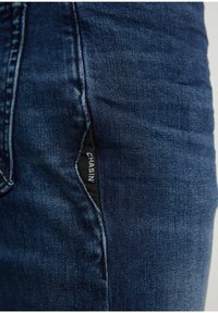 Détail d’un jean en denim bleu foncé montrant une poche latérale avec une étiquette de marque "CHASIN" cousue à l’intérieur.