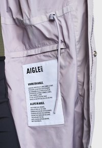 Intérieur d'une veste violet clair montrant une étiquette en tissu blanc avec la marque AIGLE et un texte bilingue en français et en anglais sur la nature et la liberté.