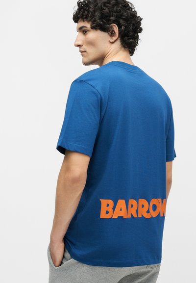 Άνδρας με κατσαρά μαλλιά που φοράει μπλε μπλουζάκι με μεγάλο πορτοκαλί κείμενο "BARROW" στο κάτω μέρος της πλάτης και γκρι παντελόνι, κοιτάζοντας πλαγίως.
