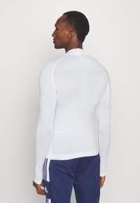 White long-sleeve athletic shirt with a smooth texture, featuring a subtle logo on the back and a fitted design. Paired with dark blue pants.