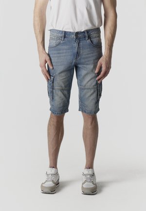 Mann trägt ausgewaschene blaue Denim-Cargoshorts und weiße Turnschuhe und steht vor einem schlichten Hintergrund.