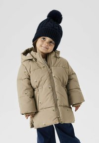 Doudoune beige avec broderie florale, fermeture éclair et capuche. Associée à un bonnet en tricot bleu marine avec un pompon sur le dessus.