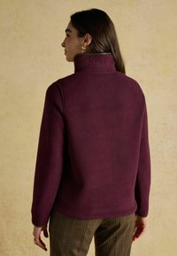 Sieviete ar gariem brūniem matiem, tērpusies augstu apkakli iespīlētu plūmju krāsas vilnas jaku un brūnām raibajām biksēm, stāv pretī gaiši brūnai fakturētai sienai.