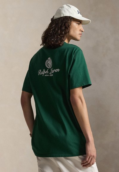 Person trägt ein dunkelgrünes Ralph-Lauren-T-Shirt mit Logo und Text auf der Rückseite, weiße Kappe und hellfarbene Hose und steht seitlich.