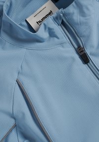 Ljusblå zip-up-jacka i texturerat tyg, med hög krage, kontrasterande grå sömmar och en märkesetikett inuti halsringningen.