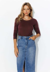 Haut long bordeaux associé à une jupe en denim taille haute avec une fente devant. Cheveux lâchés, modèle posé sur fond blanc.