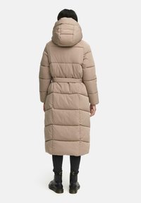 Lichtbruin gewatteerd jack met een capuchon, taille-ingeklemd en lange mouwen. Heeft verticale stiksels en een lengte tot halverwege het onderbeen. Zwarte laarzen zichtbaar.