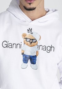 Sudadera blanca con un gráfico de un oso que lleva una camisa de patrón, con gafas y una gorra, pantalones vaqueros azules y un reloj, con el texto "Gianni Gavanagh".