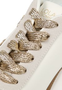 Zapatilla blanca con parte superior de ante beige, cordones trenzados metálicos dorados y suela de goma texturizada. Presenta un logo dorado en la lengüeta.