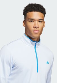 Biała sportowa koszulka z krótkim zamkiem i jasno niebieskimi akcentami, wykonana z teksturowanej tkaniny. Na lewej piersi znajduje się logo Adidas w tym samym odcieniu niebieskiego.