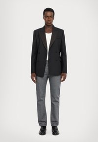 Svart dubbelknäppt blazer med satinrevers, kombinerad med en vit skjorta och grå jeans. Modellen bär svarta läderskor.