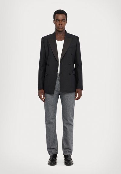 Zwarte dubbelgeknoopte blazer met satijnen revers, gecombineerd met een wit shirt en grijze jeans. Model draagt zwarte leren schoenen.
