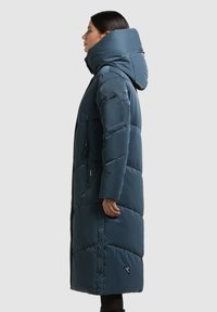 Dlouhý prošívaný kabát tmavě petrolejové barvy, vyrobený z lesklé tkaniny. Má vysoký límec, kapsy na zip a druky po stranách.