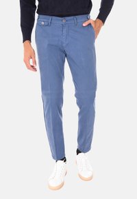 Pantaloni in cotone blu con una vestibilità slim, dotati di zip frontale, passanti per la cintura e tasche laterali. Abbinati a sneakers bianche e calzini neri.