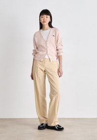 Cardigã rosa claro feito de um tecido macio, com decote em V, fechamento de botões e um pequeno logotipo. Combinado com calças bege e sapatilhas pretas.