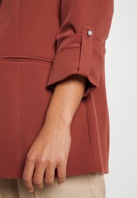 Rostfärgad, slät tygskjorta med uppvikta ärmar och pärlknotor vid manschetten. Hand vilande bredvid beige byxor.