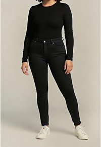 Femme portant un haut noir à manches longues et un jean skinny noir taille haute avec des baskets blanches, debout devant un fond uni.