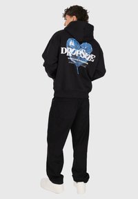 Man facing sideways wearing a black hoodie with a blue heart and white "Dropsize" text, black pants, and white sneakers on a gray background.