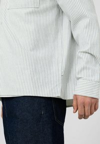 Chemise à manches longues rayée blanche et bleue, avec une poche poitrine unique, un ourlet légèrement arrondi et des poignets structurés. Portée avec un jean foncé.