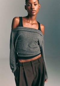 Szary sweter typu off-the-shoulder z rypsowym wykończeniem, nałożony na czarną koszulkę na ramiączkach. W połączeniu z spodniami w kolorze ciemnego brązu o wysokim stanie z guzikiem.